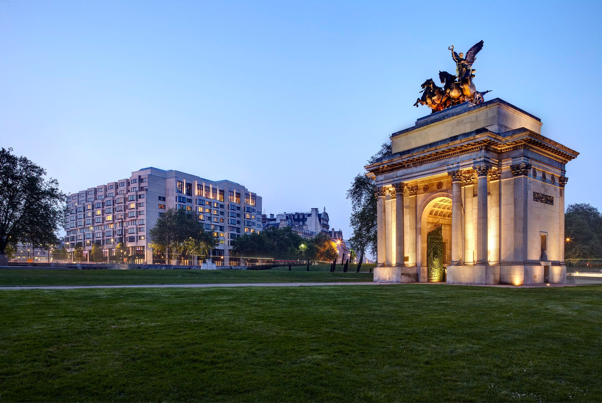 outdoor common area with green grass, an archway lit up in the evening and a hotel in the background.